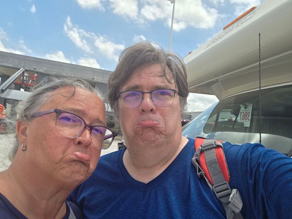 A man and a woman making sad faces while posing for a selfie outdoors, with a vehicle and a cloudy sky in the background.