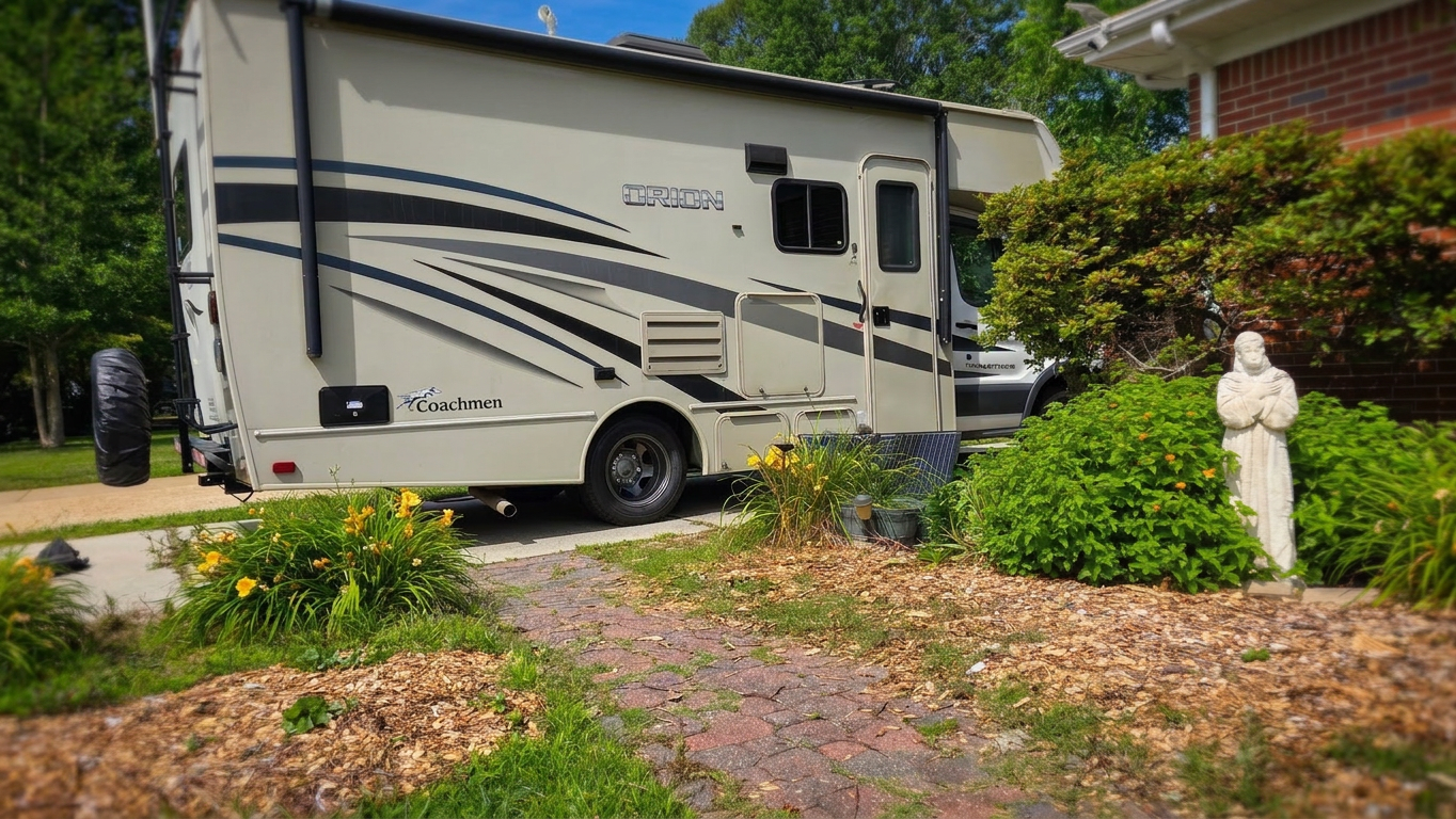 A parked RV with a beige exterior and black stripes, surrounded by green grass and flowering plants, with a stone statue in the foreground.
