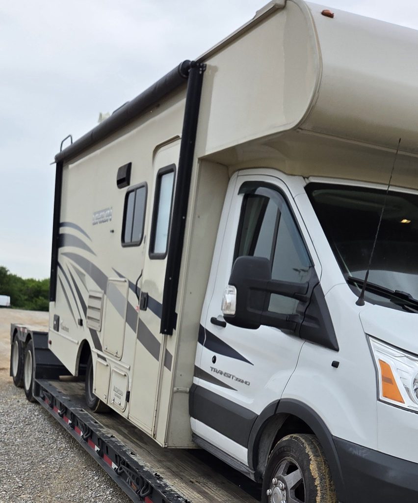 A beige and grey recreational vehicle (RV) parked on a trailer, showcasing its side view, with two windows and a front cab visible.
