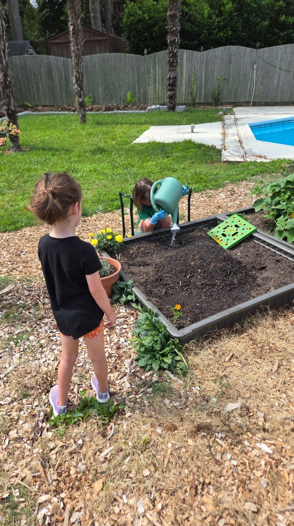 Two young children engaged in gardening; one is watering a flowerbed while the other stands nearby holding a plant pot.
