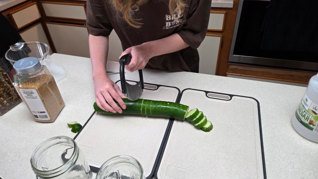 A person slicing a cucumber on a cutting board in a kitchen, with various ingredients and jars in the background.