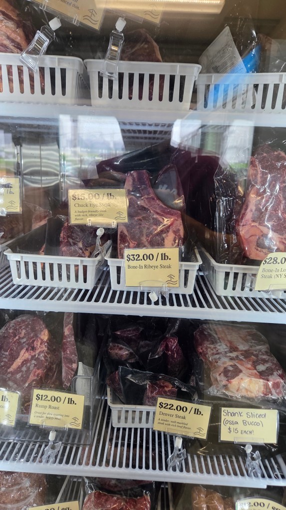 Display case with various cuts of beef, including Bone-In Ribeye Steak priced at $32.00/lb, Chuck Eye Steak at $16.00/lb, Rump Roast at $12.00/lb, Denver Steak at $22.00/lb, and Shank Sliced (Ossa Bucco) at $15 each. Labels provide pricing and descriptions for each cut.